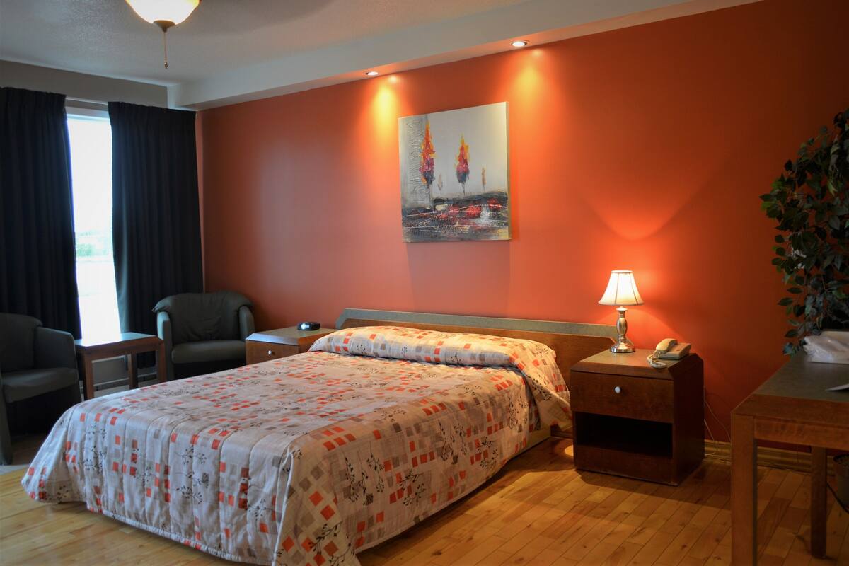 A hotel room with a bed, a desk, a chair, a nightstand with a lamp, and a painting on the wall.