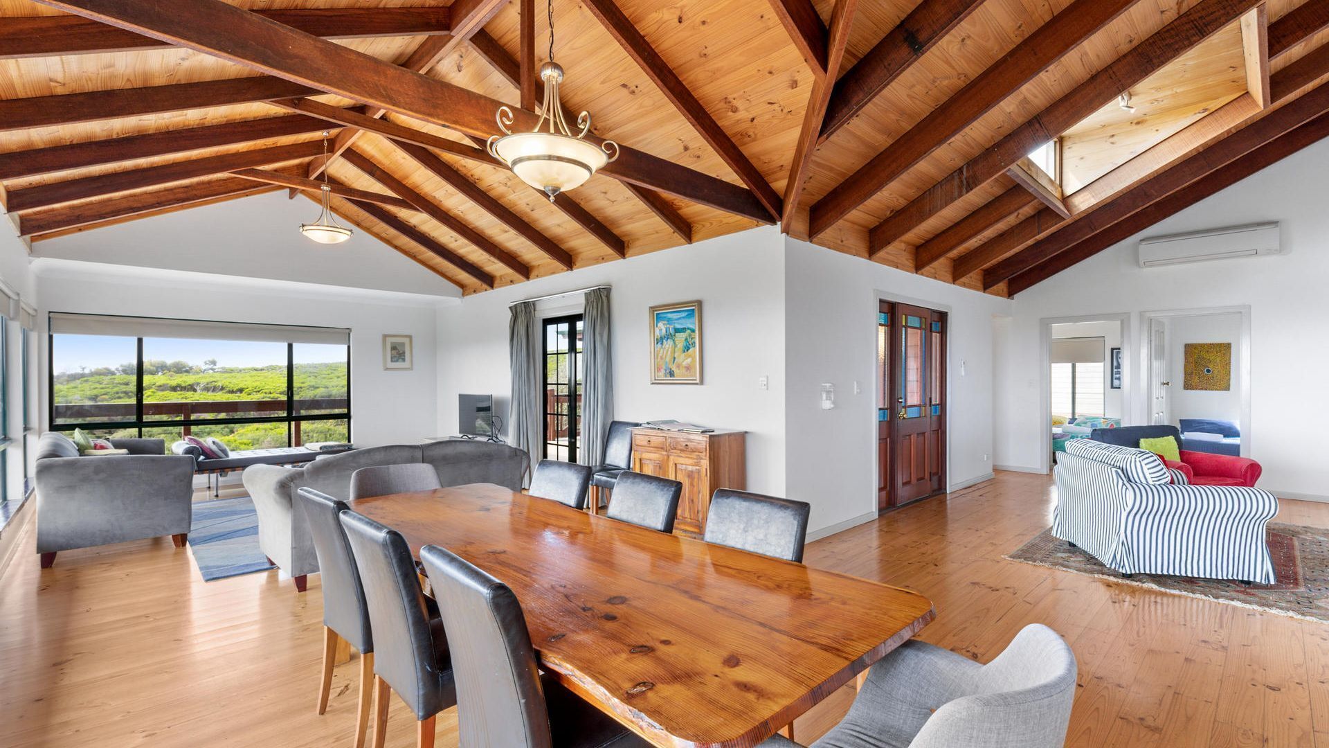 A spacious dining area with a wooden table and chairs, a large window with a view of greenery, and a high ceiling with exposed beams.