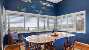 Un coin repas avec une table ronde en bois et des chaises rembourrées bleues, de grandes fenêtres avec des volets blancs, et une fresque murale représentant des poissons.