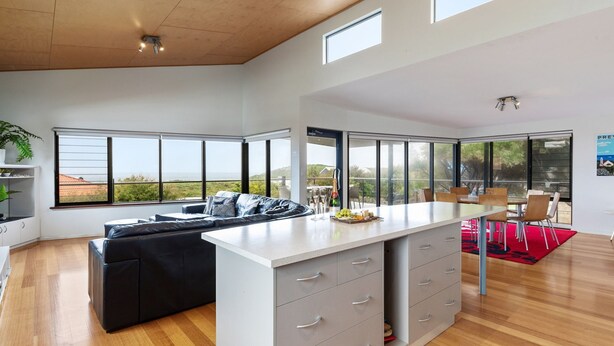 A spacious living area with a large window, a black sofa, a kitchen island, and a dining area with chairs. - Beachside at Margaret River (Prevelly) - Modern beach house, ocean views, spacious interior, reverse (PREVELLY)