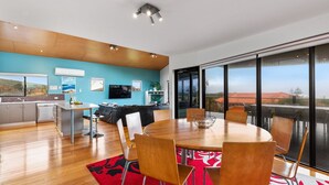A modern kitchen with a wooden ceiling, a dining area with a round table and chairs, and a large window with a view of a red-roofed building. - Beachside at Margaret River (Prevelly) - Modern beach house, ocean views, spacious interior, reverse (PREVELLY)