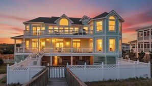 Una amplia casa de varios pisos frente a la playa, con un porche cubierto y una terraza de madera que conduce a la entrada.