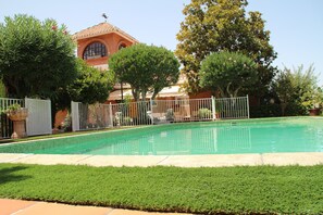 A swimming pool with a tiled deck, surrounded by a white fence, and a building with a red-tiled roof in the background.