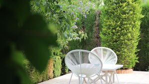 Un patio al aire libre con dos sillas blancas y una mesa, rodeado de follaje cuidado y un seto.