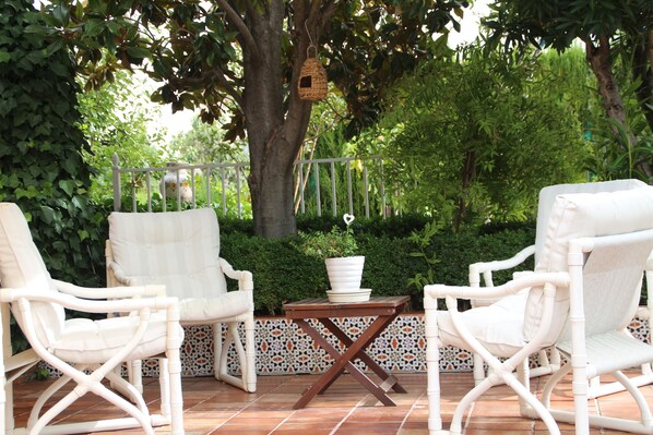 An outdoor seating area with wicker chairs and a wooden table, surrounded by a tiled patio and lush greenery.