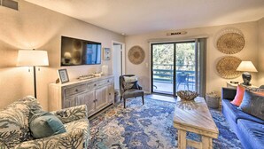A living room with a sofa, armchair, and coffee table, a television, and a large window with a view of trees. - Half-Mi to Beach: Hilton Head Condo w/ Lagoon View (Hilton Head)