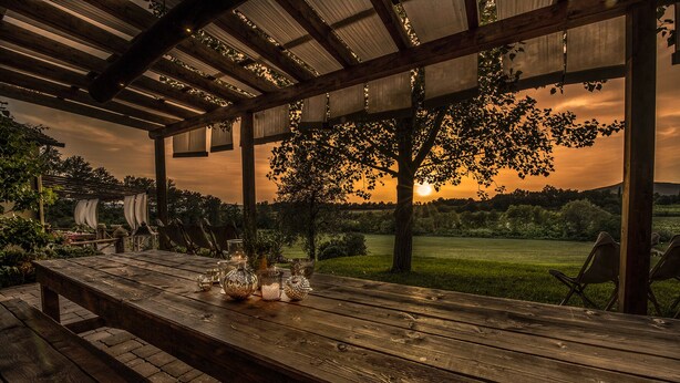 A wooden table set for dining with candles and glassware, under a pergola with a view of a sunset over a grassy field. - Romignano wine farm. Your villa in TUSCANY. (Traiana)