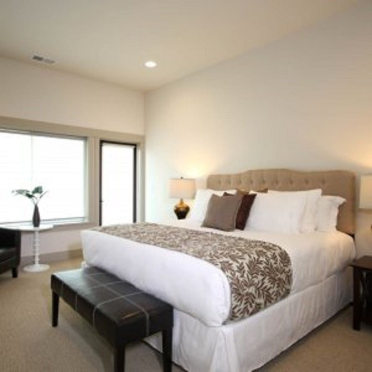 A hotel room with a large bed, a bedside table with a lamp, a small table with a plant, and a window with blinds.