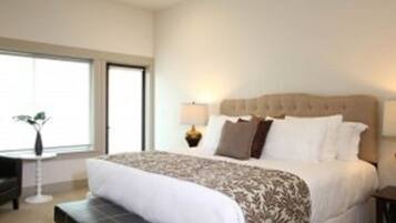 A hotel room with a large bed, a bedside table with a lamp, a small table with a plant, and a window with blinds.