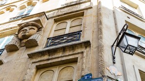 A building facade with architectural details, including a carved stone head, a balcony with a wrought iron railing, and street signs reading "Place Ducaire" and "Rue Ducaire". - Classic Style by Rue Montorgueil, by Veeve (Paris)