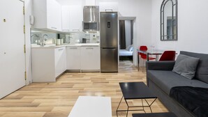 A modern kitchen with white cabinets, a stainless steel refrigerator, and a microwave. There is a dining area with a red chair and a white table. A gray sofa is in the living area. - Modern Madrid Downtown Puerta Del Sol M (PRE2B) (Madrid)