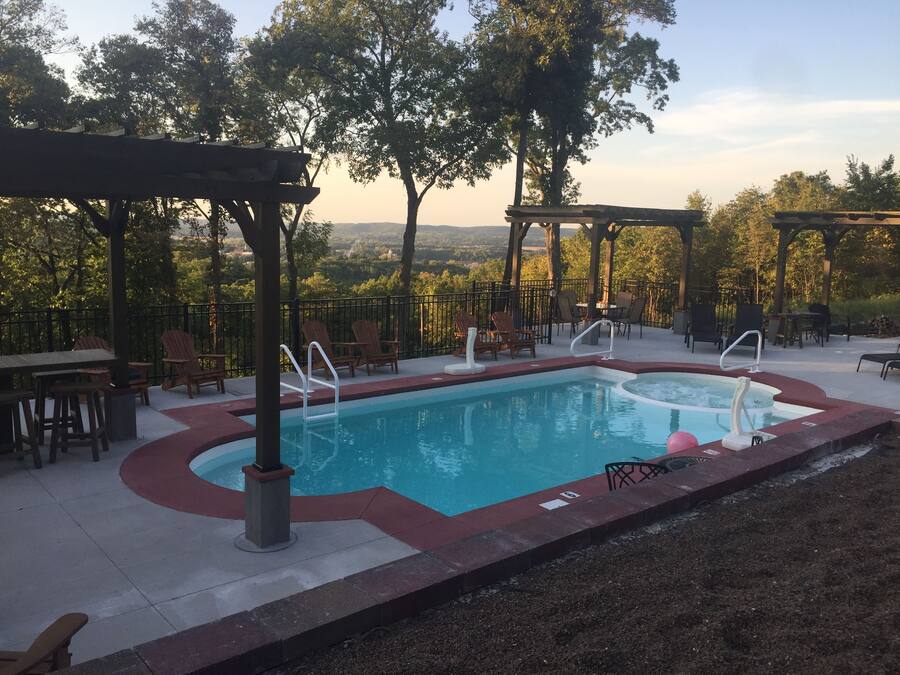 A swimming pool with a red border, surrounded by lounge chairs and tables, set against a backdrop of trees and a distant view.