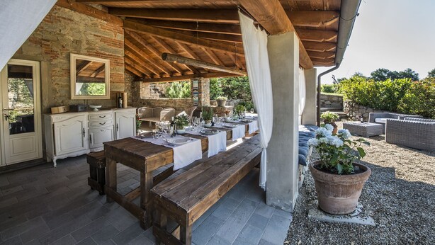An outdoor dining area with a rustic wooden table set for a meal, a stone wall, and a seating area with wicker furniture. - CHARMING VILLA IN THE HEART OF CHIANTI (terranuova bracciolini)