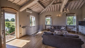 A spacious living room with a high ceiling, wooden beams, a grey sofa set, a chandelier, and large windows offering a view of greenery outside. - CHARMING VILLA IN THE HEART OF CHIANTI (terranuova bracciolini)