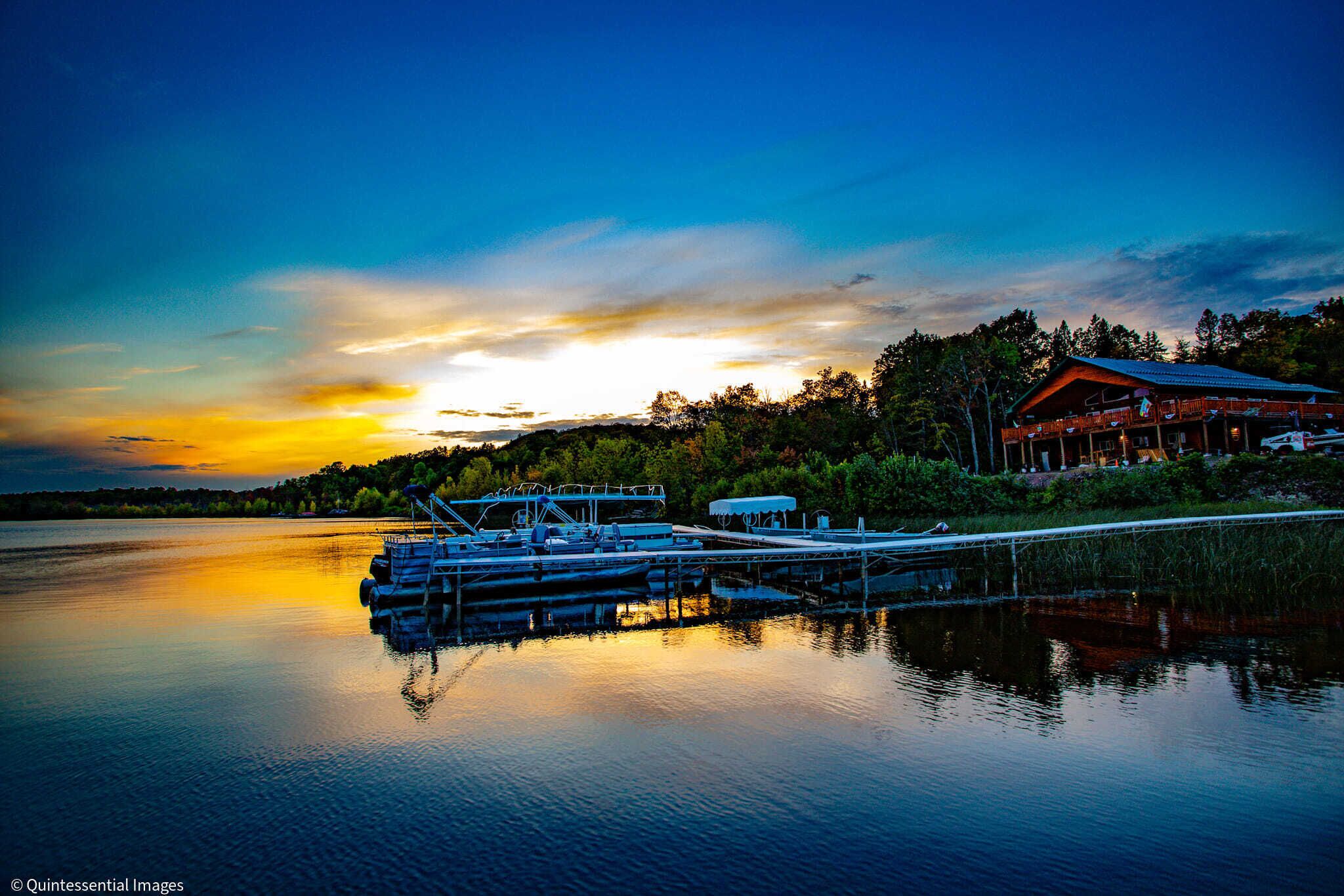 A lakeside cabin with docked boats, surrounded by trees and a sunset.