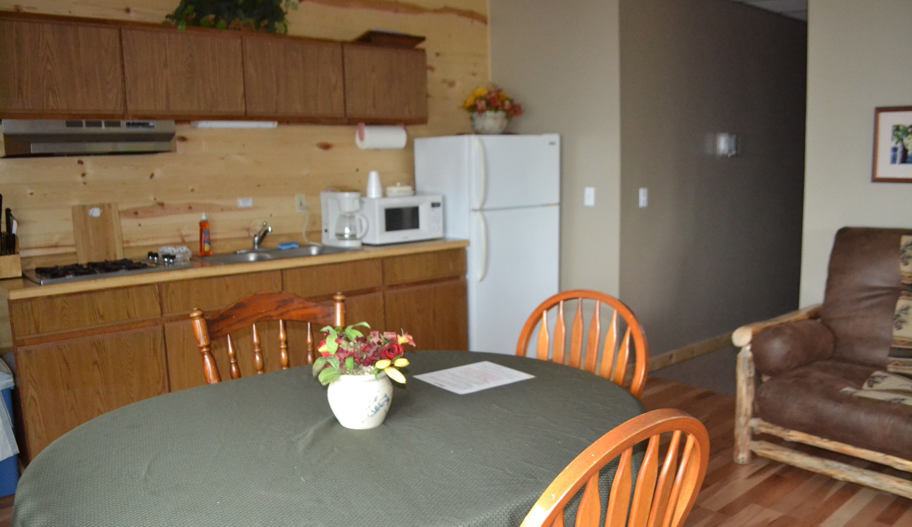 A kitchen with wooden cabinets, a microwave, and a refrigerator. There is a dining table with chairs and a vase with flowers.