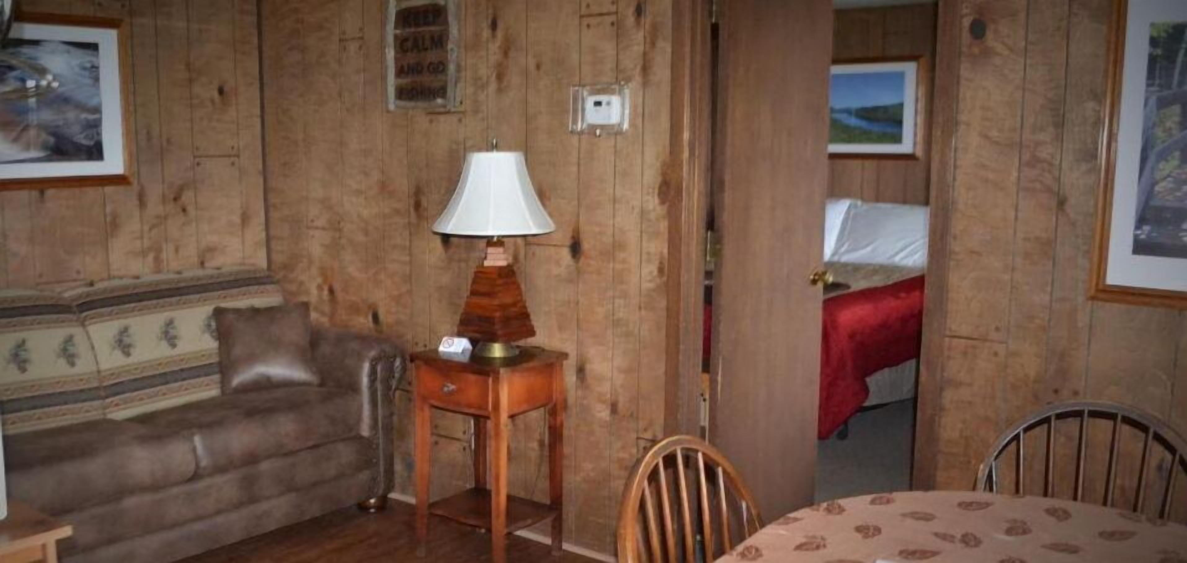 A wooden-furnished room with a sofa, a table with a lamp, and a bed with a red blanket.