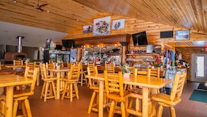 A rustic wooden interior with multiple wooden tables and chairs, a bar area with various bottles, and a fireplace. - AJ's Lodge and Oven (Bergland)