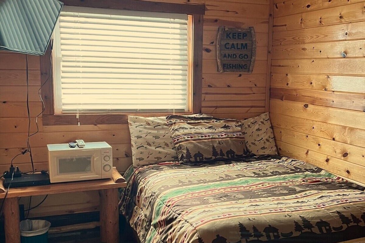 Une chambre confortable de type chalet, avec un lit, une petite table équipée d’un micro-ondes, une fenêtre avec des stores et un panneau affiché sur le mur.
