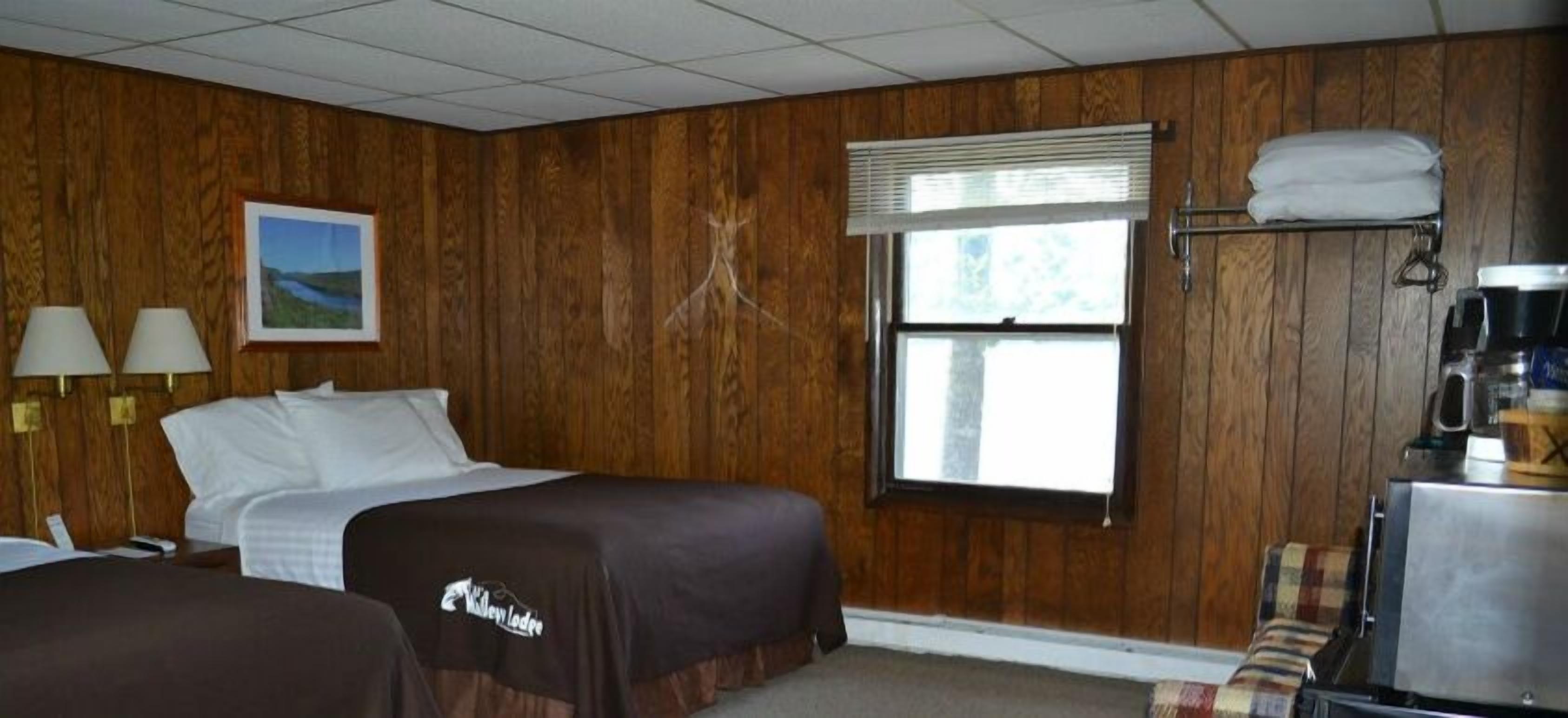 Una habitación de hotel con paneles de madera, una cama, un escritorio, una silla, un ventanal con persianas y una cafetera.