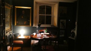 Un comedor tenuemente iluminado con una mesa dispuesta para dos, una ventana con vistas y un cuadro en la pared.