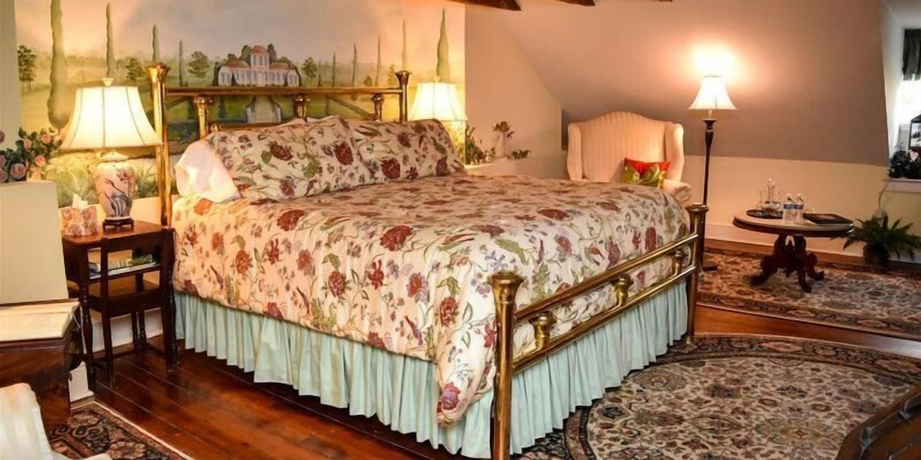 A bedroom with a floral bedspread, a wooden headboard, a nightstand, a lamp, and a view of a landscape painting.