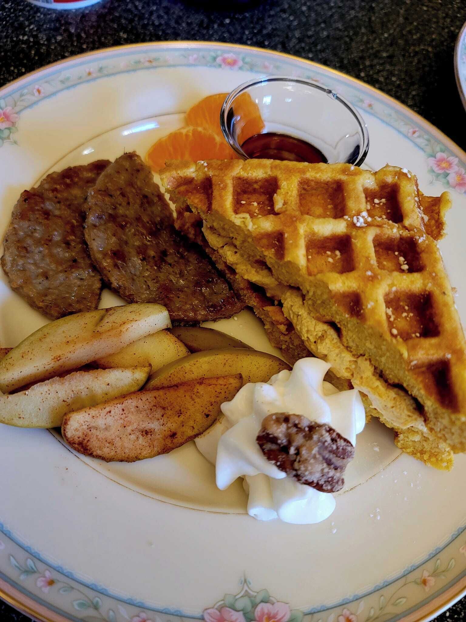 A plate with a waffle, meat patties, fries, and a small bowl of sauce.