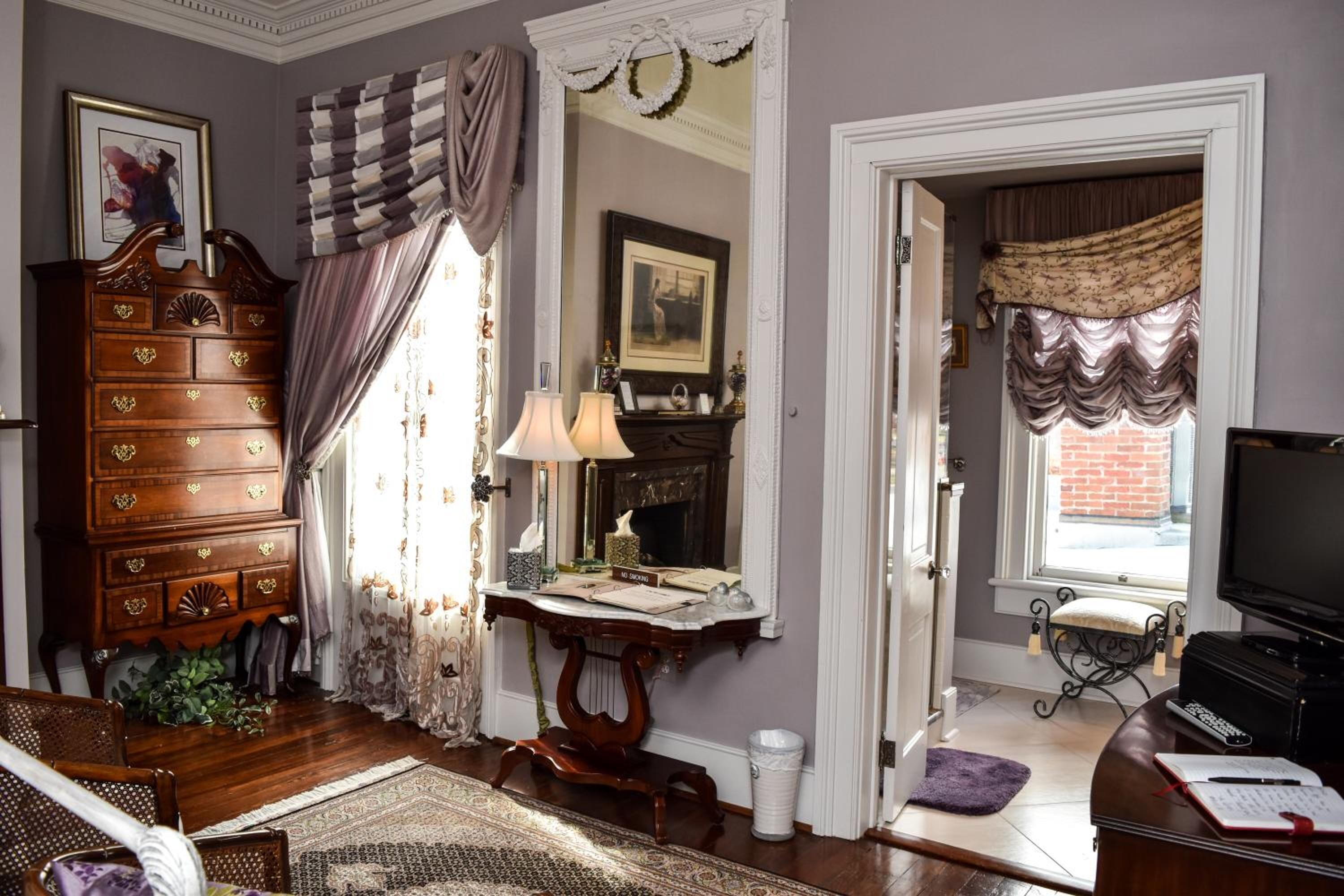 A traditional room with a wooden dresser, a desk with a lamp, a television, and a window with curtains.