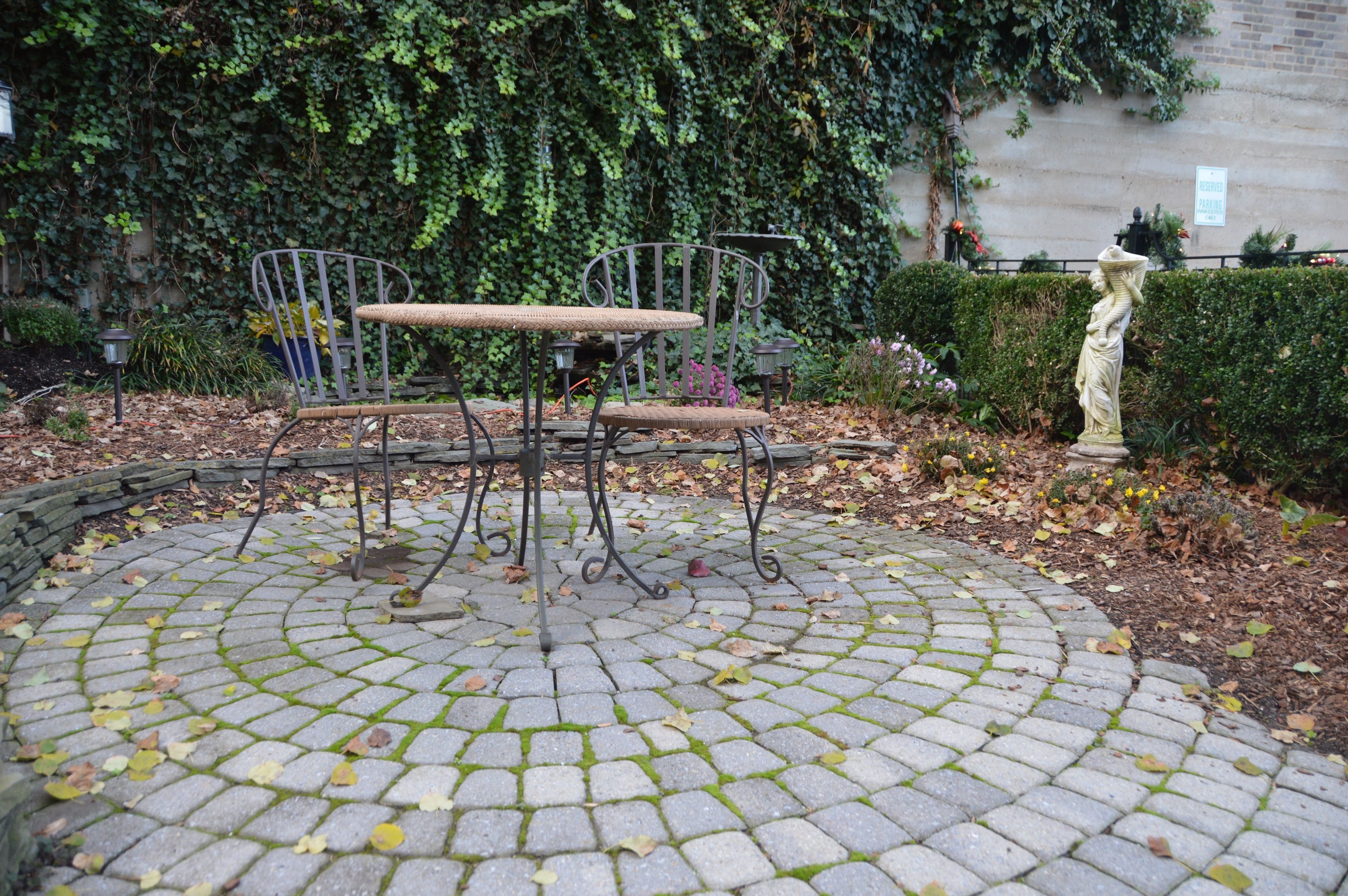 A circular stone patio with a metal table and chairs, surrounded by a hedge and a statue.