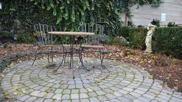 A circular stone patio with a metal table and chairs, surrounded by a hedge and a statue.