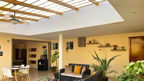 A spacious living area with a high ceiling, a dining table with chairs, a sofa with cushions, and several potted plants.