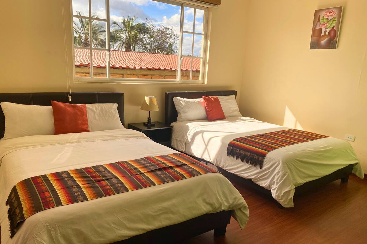 A hotel room with two beds, a window with a view of palm trees and a red-roofed building, and a painting on the wall.