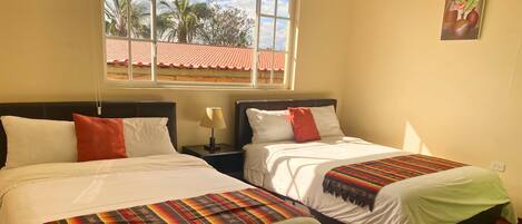 A hotel room with two beds, a window with a view of palm trees and a red-roofed building, and a painting on the wall.