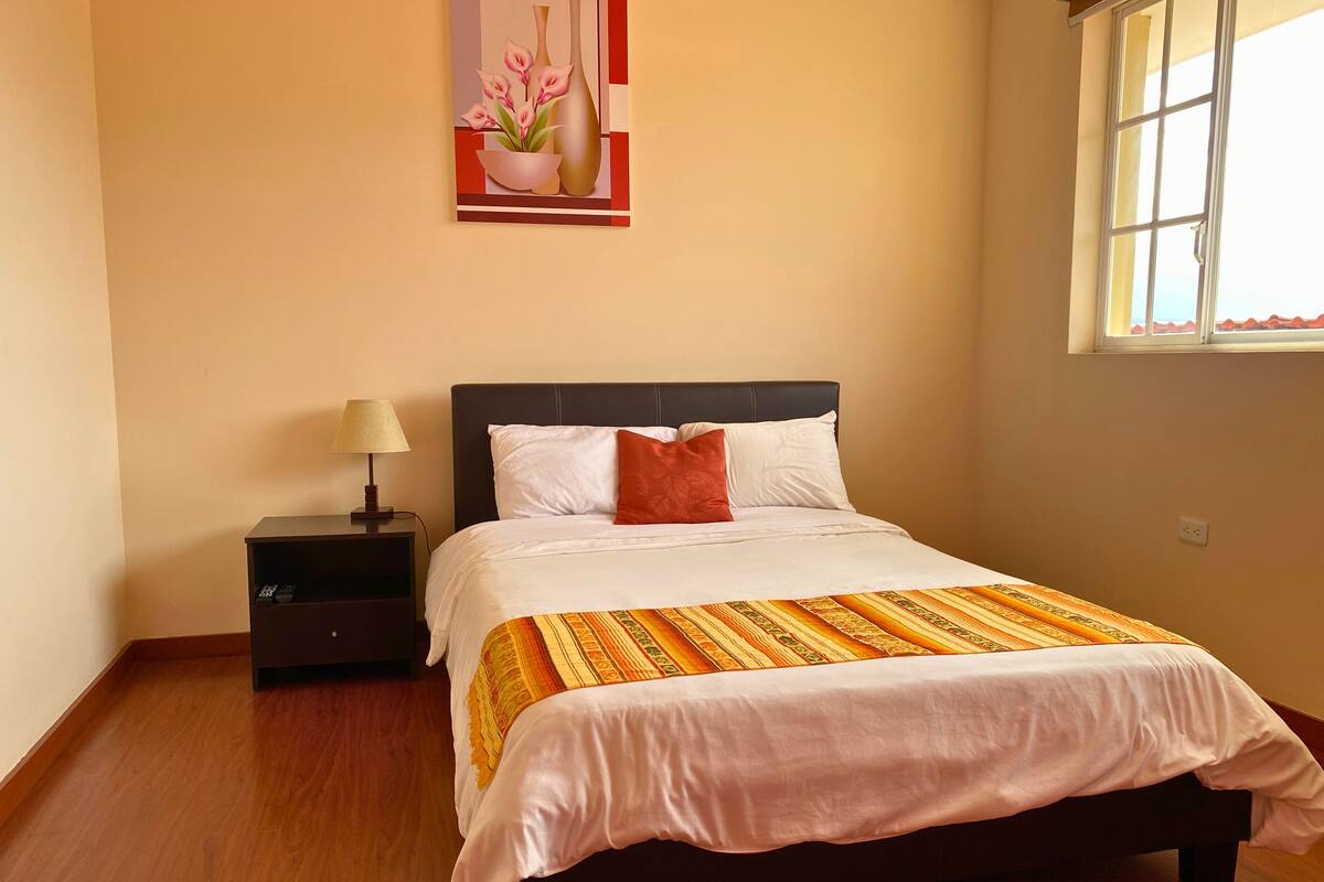 A neatly made bed with a white comforter and a patterned throw, a bedside table with a lamp, and a painting of flowers on the wall.