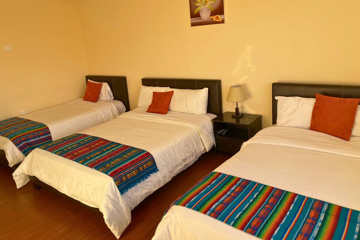 A hotel room with three beds, each with white bedding and colorful blankets. There is a nightstand with a lamp between the beds.