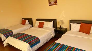 Habitación de hotel con tres camas, cada una con ropa de cama blanca y mantas coloridas. Hay una mesita de noche con una lámpara entre las camas.