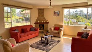A living room with a stone fireplace, two sofas, a coffee table, and large windows offering a view of a garden and buildings.