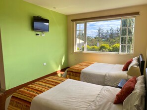A room with two beds, a television mounted on the wall, and a window with a view of trees and buildings. - Las Hortensia's Casa De Huéspedes (Tababela)