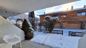 Ein überdachter Außenbereich mit einer Bank, schneebedeckten Pflanzen und einem Holzgebäude im Hintergrund.