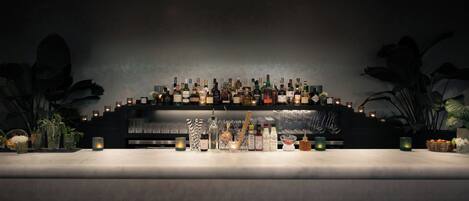 Zona de bar con una barra larga, varias botellas, vasos y algunas plantas en macetas.