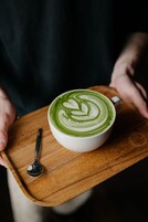 Iemand houdt een houten dienblad vast met een kop groene koffie en een lepel.