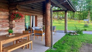 A wooden cabin with a patio area featuring a wooden table and chairs, a hanging flower arrangement, and a view of the surrounding greenery.