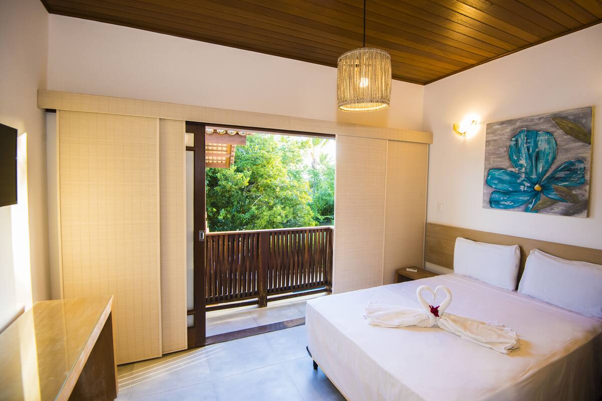 A hotel room with a bed, a wooden ceiling, a painting, and a view of greenery outside.