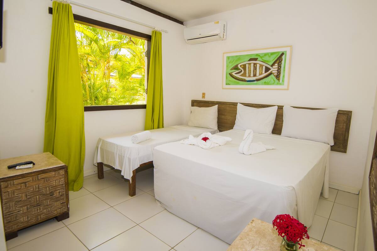 A hotel room with two beds, a small table with a vase of red flowers, a nightstand, and a window with green curtains.