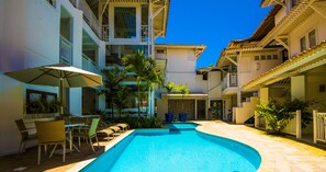 Une piscine entourée de bâtiments dotés de balcons et agrémentée de plantes en pot, avec des chaises longues et des parasols à proximité.