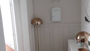 Une pièce avec une porte blanche, un lampadaire, une petite table blanche avec une lampe et une boîte aux lettres fixée au mur.