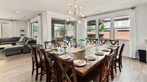 A dining area with a wooden table set for six, surrounded by wooden chairs, with a chandelier above and large windows offering a view of the outdoors. - 94 PV, Luxury Home with Private Pool & Hot Tub, Fire Pit, Huge Kitchen, and More! (Santa Clara)