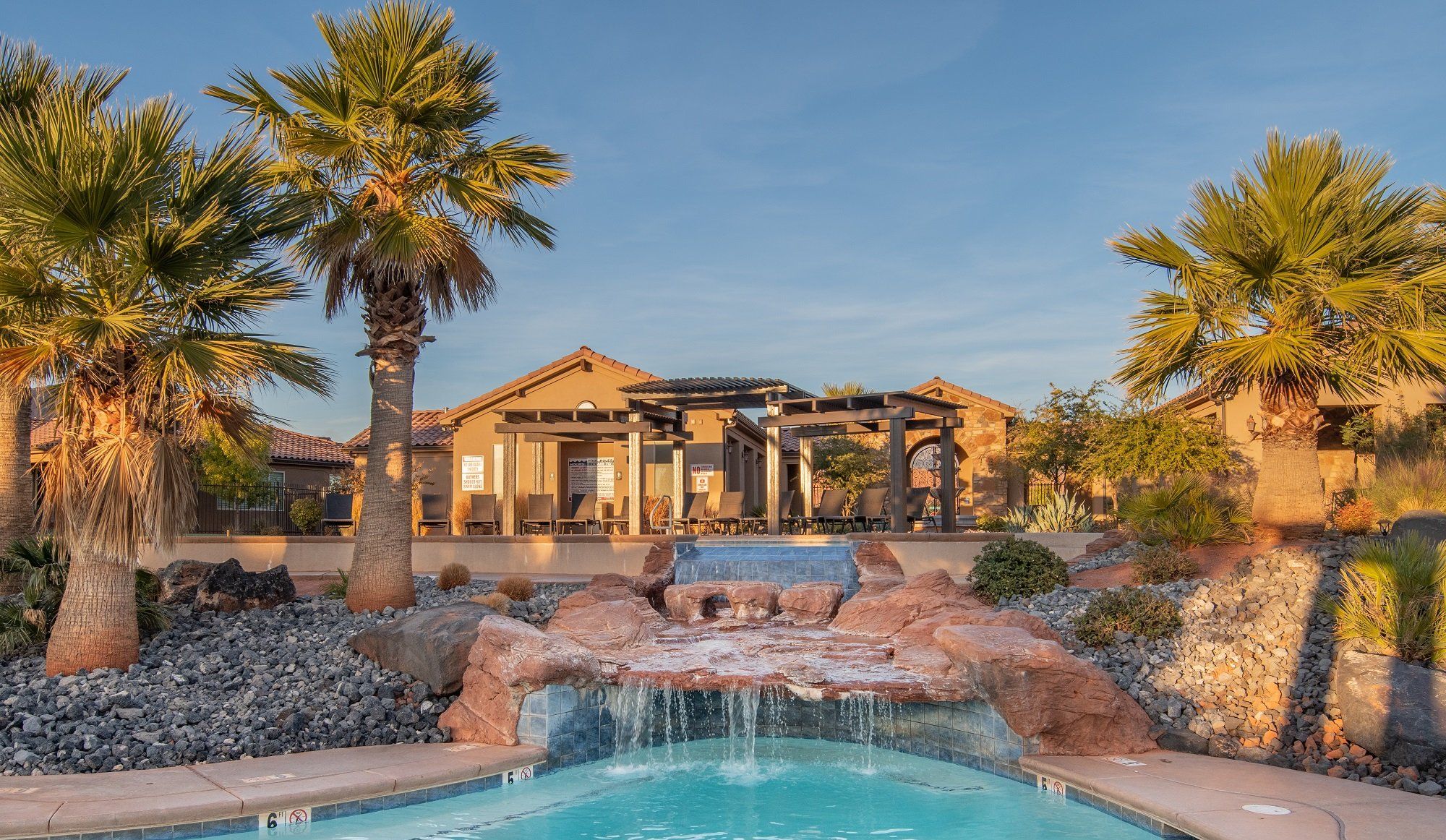 A pool with a waterfall feature, surrounded by rocks and palm trees, with a building in the background.
