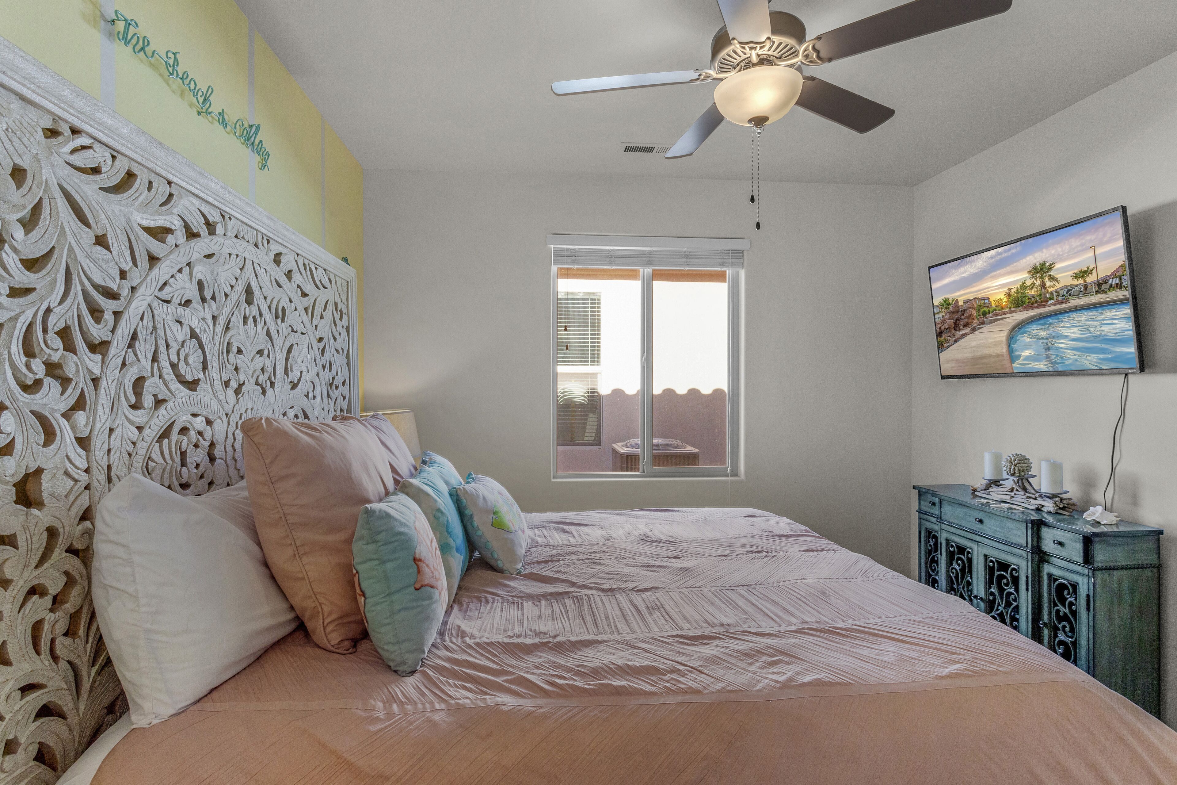 Une chambre à coucher avec une tête de lit décorative, un lit avec plusieurs oreillers, un ventilateur de plafond, une fenêtre avec des stores et un téléviseur fixé au mur.