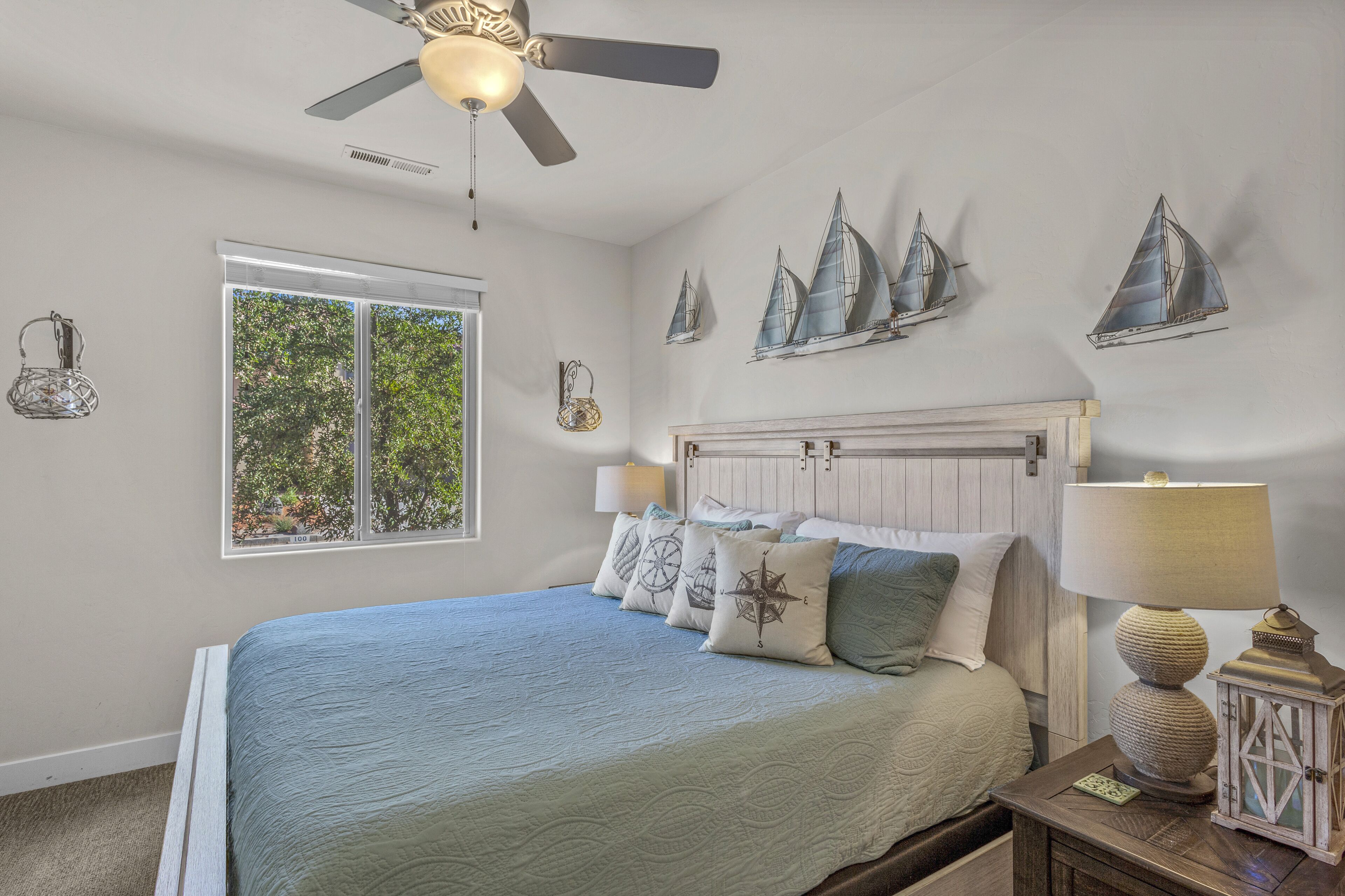 Une chambre avec un lit, un ventilateur de plafond, une fenêtre donnant sur un beau paysage et des tableaux représentant des voiliers.
